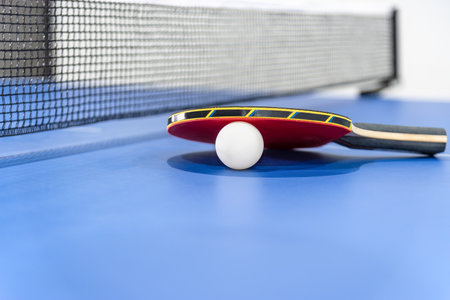 Closeup red table tennis racket and a white ball on the blue table with a black net, Table tennis paddle is a sports competition equipment indoor activity and exercise for concept backgroundの写真素材