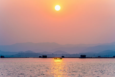 Beautiful nature landscape silhouette fisherman driving a boat to home under the sun, Golden light on the red orange sky at sunset local fishing rural lifestyle at Krasiao Dam, Suphan Buri, Thailandの写真素材