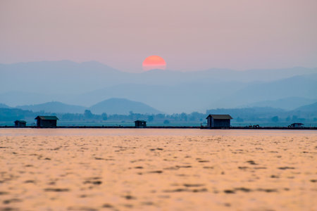 Beautiful nature landscape orange sun in the red sky sunlight over water lake blue mountain background at sunset, silhouette fish farm cages rural lifestyle at Krasiao Dam, Suphan Buri, Thailandの写真素材