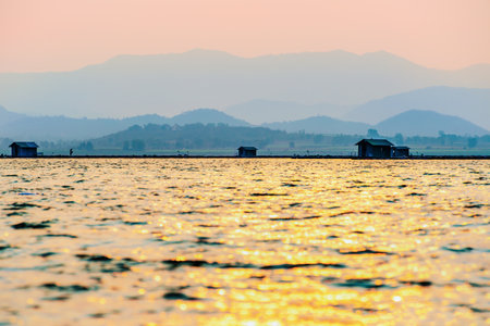 Beautiful nature landscape silhouette fisherman and fish farming cages, golden sunlight reflecting on the water surface at sunset, Local fishing rural lifestyle at Krasiao Dam, Suphan Buri, Thailandの写真素材
