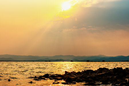 Beautiful nature landscape sun shines bright golden in the cloud reflect yellow light on the sky pile of rock in the water of a tropical lake mountain background at Krasiao Dam, Suphan Buri, Thailandの写真素材