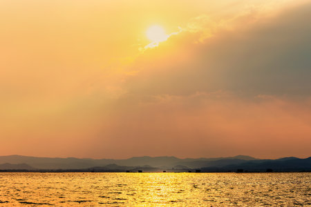 Beautiful nature landscape sun shines bright golden in the cloud reflect yellow light on the sky and water surface tropical lake in summer mountain background at Krasiao Dam, Suphan Buri, Thailandの写真素材