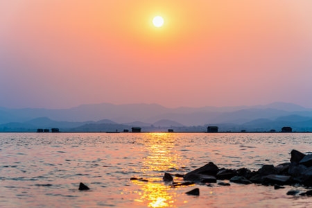 Beautiful nature landscape silhouette fisherman in a boat fishing and fish farming cages yellow sun in the red orange sky at sunset, Local fishing rural lifestyle at Krasiao Dam, Suphan Buri, Thailandの写真素材