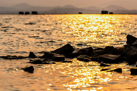 Beautiful nature landscape of bright golden sunlight reflects the yellow glow on the ripples pile of rock in the water of a tropical lake mountain background at Krasiao Dam, Suphan Buri, Thailandの写真素材