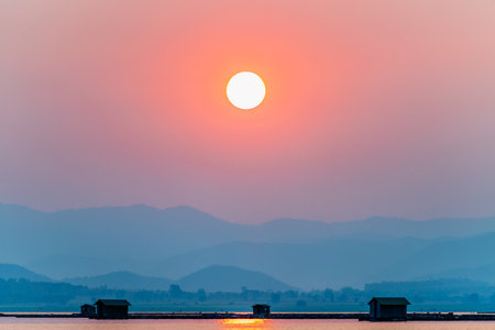 Beautiful nature landscape orange sun in the red sky sunlight over water lake blue mountain background at sunset, silhouette fish farm cages rural lifestyle at Krasiao Dam, Suphan Buri, Thailandの写真素材