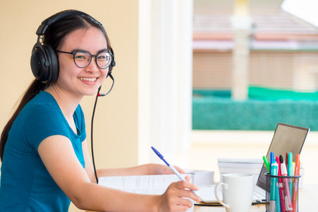 Young student Asian teenage girl with headphones and glasses is smiling happy looking at laptop computer online learning from school. Distance education class of the university video call at a homeの写真素材