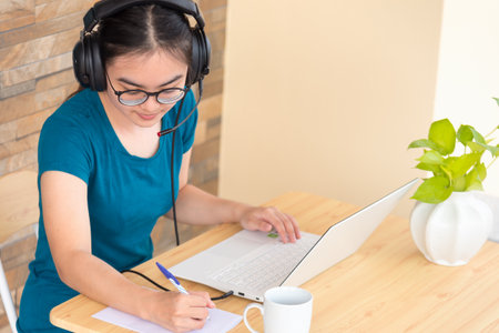Young student Asian teenage girl with headphones and glasses is smiling happy looking at laptop computer online learning from school. Distance education class of the university video call at a homeの写真素材