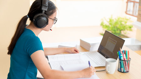 Young student Asian teenage girl with headphones and glasses is smiling happy looking at laptop computer online learning from school. Distance education class of the university video call at a homeの写真素材