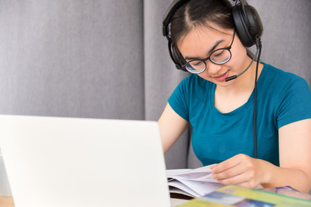 Young student Asian teenage girl with headphones and glasses is smiling happy looking at laptop computer online learning from school. Distance education class of the university video call at a homeの写真素材