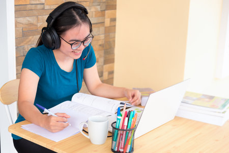 Young student Asian teenage girl with headphones and glasses is smiling happy looking at laptop computer online learning from school. Distance education class of the university video call at a homeの写真素材