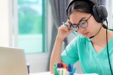 Asian woman student teenage girl with glasses headphones sitting looking serious reading a book worry using laptop computer on table learning online study. Education from a class of university at homeの写真素材