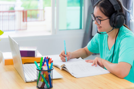 Asian young woman student with glasses headphones girl study happy writing note on a book looking video conference laptop computer university class online internet learning distance education at homeの写真素材