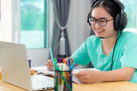 Asian young woman student with glasses headphones girl study happy writing note on a book looking video conference laptop computer university class online internet learning distance education at homeの写真素材