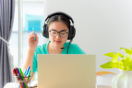Asian young woman student with glasses headphones girl study serious writing note on book looking video conference laptop computer university class online internet learning distance education at homeの写真素材