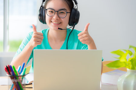 Portrait Asian young woman student glasses headphones happy study smile looking two thumbs up online class college learning internet education teenage girl work distance on a laptop computer from homeの写真素材