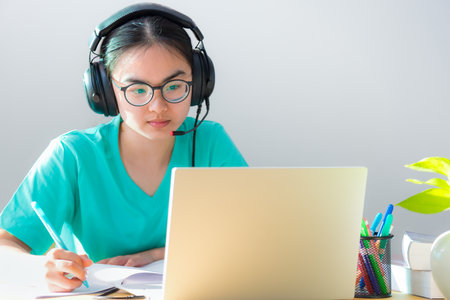 Asian young woman student with glasses headphones study serious writing note on a book looking video conference on laptop computer university class online internet learning distance education at homeの写真素材