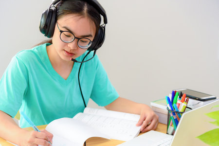 Asian woman student teenage girl with glasses headphones sitting happy smile looking writing notes at book laptop computer on table learning online study education from the class of university at homeの写真素材