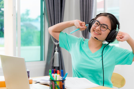 Portrait Asian young woman student glasses headphones stretch oneself from study online class college learning internet education smile looking up teenage girl work distance on laptop computer at homeの写真素材
