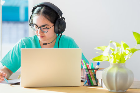 Asian woman student teenage girl with glasses headphones sitting happy smile looking writing notes at book laptop computer on table learning online study education from the class of university at homeの写真素材
