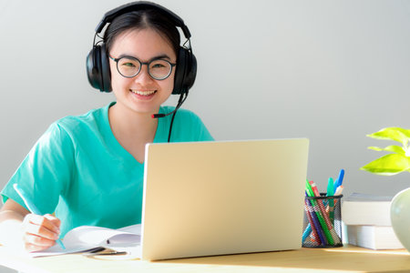 Portrait young Asian female student with headphones glasses smile looking up a note on the book a teenage girl is happy to university internet distance study online class on a laptop computer at homeの写真素材