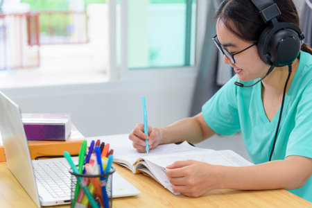 Asian woman student teenage girl with glasses headphones sitting happy smile looking writing notes at book laptop computer on table learning online study education from the class of university at homeの写真素材