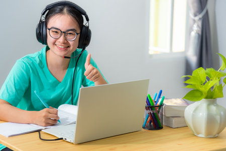 Portrait young Asian female student with headphones glasses smile looking and thumb up a teenage girl is happy to university internet distance study online class works on a laptop computer from homeの写真素材