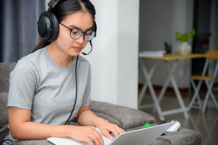 Asian young woman student with headphones sitting on the sofa look at laptop happy study online class college learning internet education, Teenage girl work distance on a laptop computer from homeの写真素材