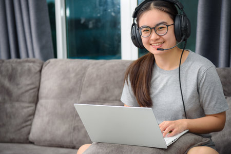 Asian young woman student with headphones sitting on the sofa smile looking up happy study online class college learning internet education, Teenage girl work distance on a laptop computer from homeの写真素材