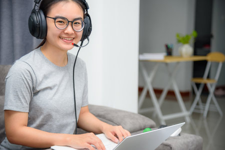 Asian young woman student with headphones sitting on the sofa smile looking up happy study online class college learning internet education, Teenage girl work distance on a laptop computer from homeの写真素材