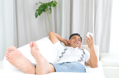 Smartphone mobile apps concept. Malay guy using smart phone. Handsome asian man relaxed and lying on sofa indoor.の写真素材