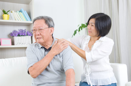 Asian old man shoulder pain, sitting on sofa , daughter massaging father shoulder. Chinese family, senior retiree indoors living lifestyle at home.の写真素材