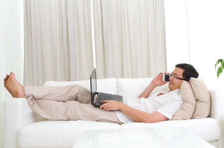 Smartphone mobile apps concept. Guy using smart phone and laptop to perform internet banking. Handsome asian man relaxed and lying on sofa indoor.の写真素材