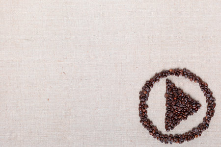 Bunch of roasted coffee beans forming play symbol enclosed in circle on creamy linen canvas, arranged bottom rightの写真素材