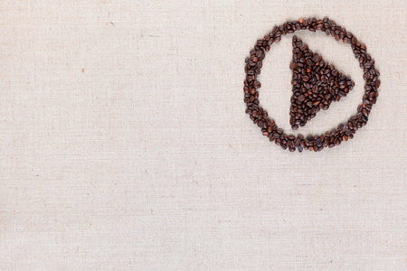Bunch of roasted coffee beans forming play symbol enclosed in circle on creamy linen canvas, arranged top rightの写真素材