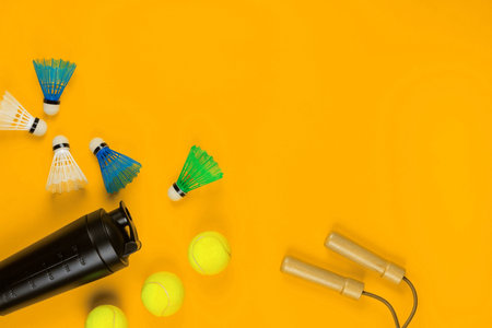 A pile of sport objects like a jump rope, shuttlecocks, water bottle, tennis balls all on yellow...
