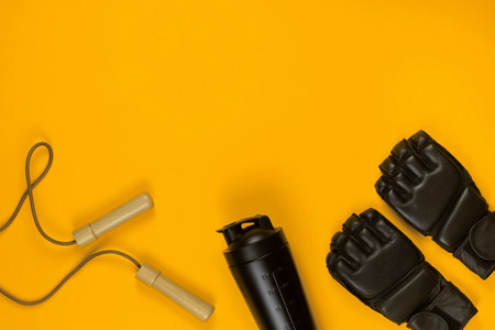 A pair of black fighting gloves next to a water bottle and a jump rope, on yellow background, shot...