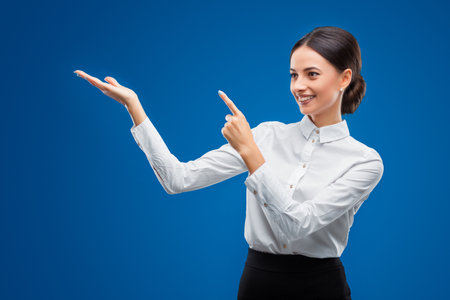 Beautiful, young businesswoman smiling and pointing towards top left, isolated on blue background.の写真素材