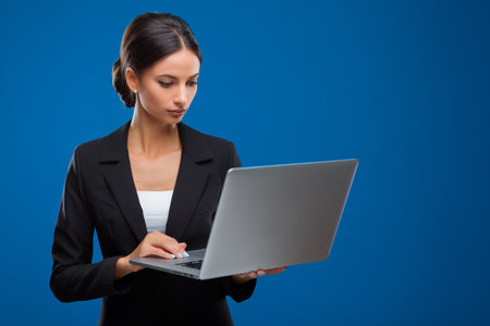 Portrait of a young and beautiful businesswoman holding and looking at a gray laptop, isolated on blue background.の写真素材