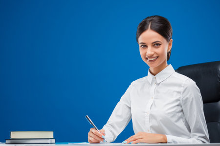 Beautiful, young businesswoman sitting at her desk and writing business concepts while smiling towards the camera, isolated on blue background.の写真素材