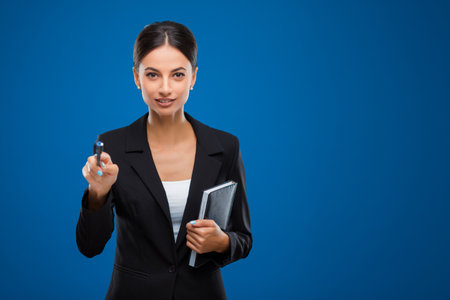 Portrait of a young and beautiful businesswoman holding a black notebook and pointing with a pen towards the camera, isolated on blue background.の写真素材