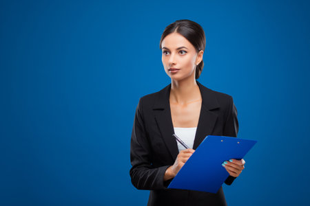 Portrait of a young and beautiful businesswoman holding a blue clipboard and a pen, isolated on blue background.の写真素材