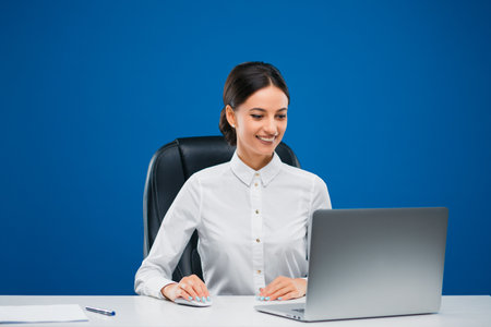 Beautiful, young businesswoman sitting at her desk and happily working at the laptop, isolated on blue background.の写真素材