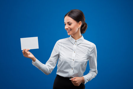 Beautiful, young businesswoman happily looking at a small blank sheet of paper which shes holding to her right, isolated on blue background.の写真素材