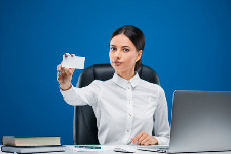 Beautiful, young businesswoman sitting at her desk before a laptop, showing a blank slip of paper, isolated on blue background.の写真素材