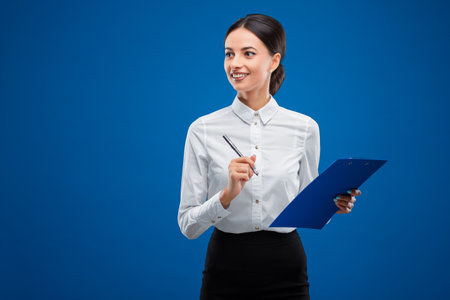 Beautiful, young businesswoman looking happily towards the left while writing on her blue clipboard, isolated on blue background.の写真素材