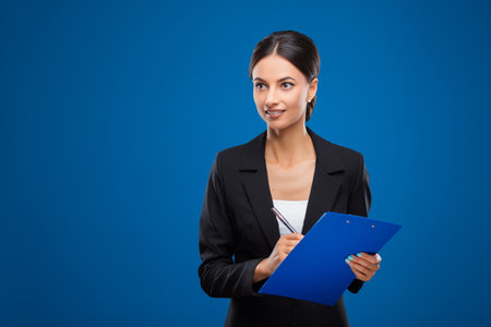 Portrait of a young and beautiful businesswoman thinking what to write on her blue clipboard, isolated on blue background.の写真素材