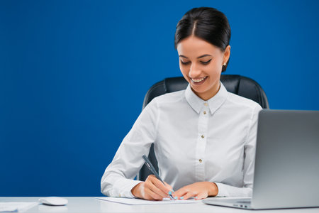 Beautiful, young businesswoman sitting at her desk before a laptop writing with enthusiasm her ideas on a sheet of paper, isolated on blue background.の写真素材