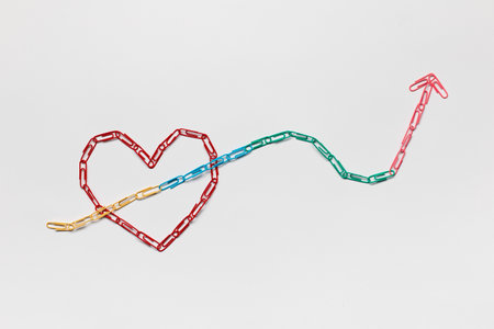 An arrow made from colored paperclips pointing upwards passing through a heart on a white background, shot from above.の写真素材