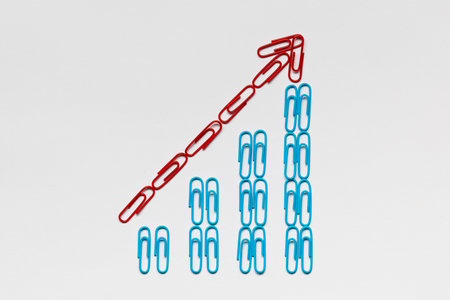 A red arrow pointing upwards siting on a stairwell made from blue paperclips on white background,...