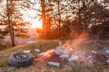 A car wheel next to a burning pile of leaves surrounded bu plastic bottles in the forest at sunrise.の写真素材